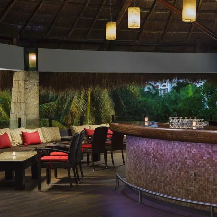 inviting interior of Papos Bar at The Reef Playacar, featuring a well-stocked bar, cozy seating with red cushions, and a thatched roof.