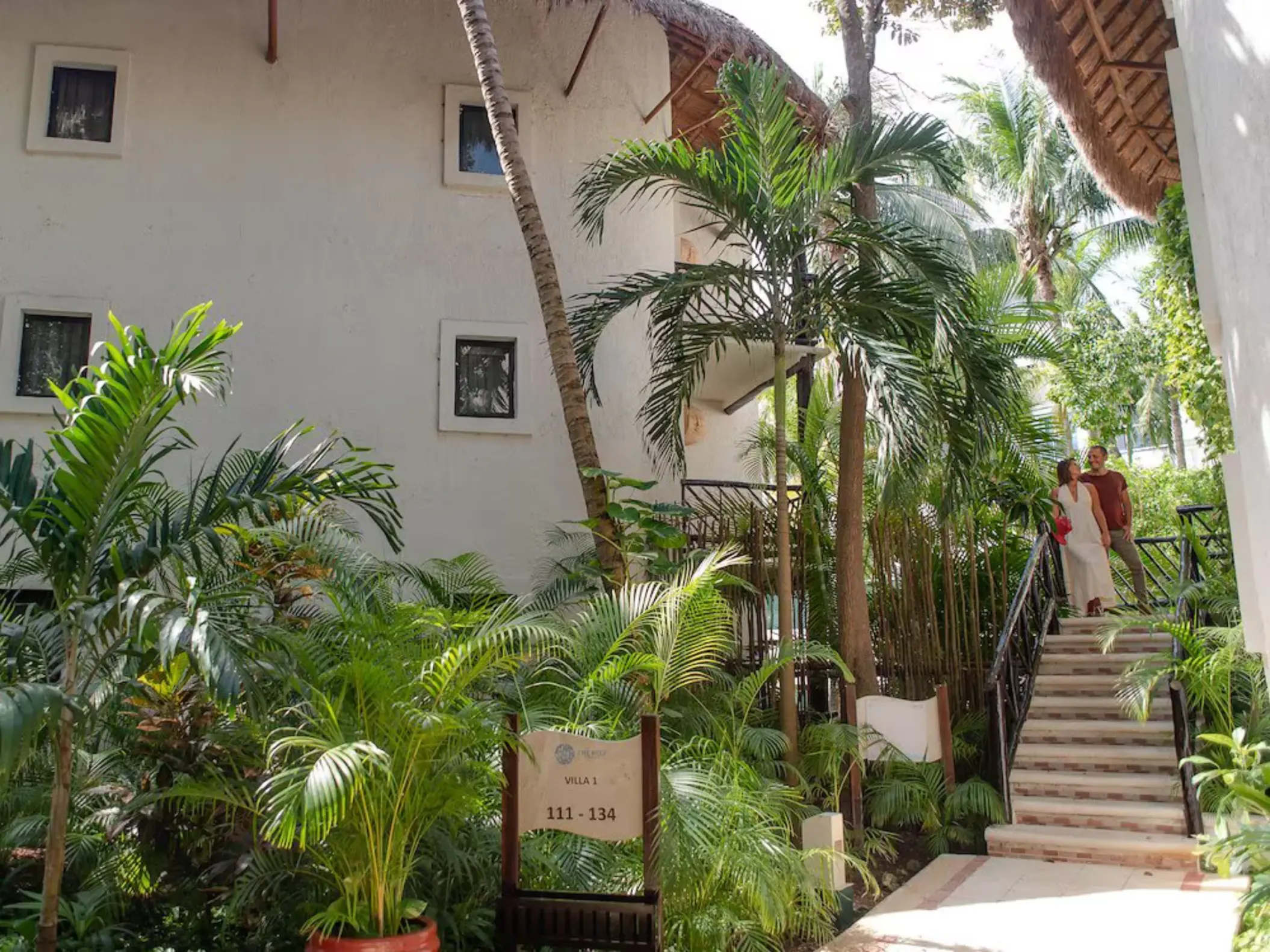 The lush tropical grounds of The Reef Playacar resort, showing white accommodation buildings, palm trees, and a couple on stairs.