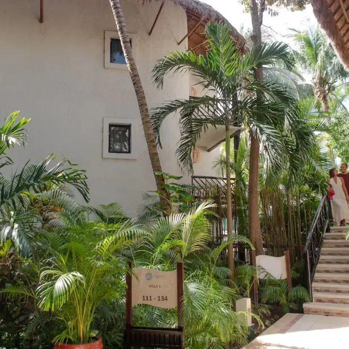The lush tropical grounds of The Reef Playacar resort, showing white accommodation buildings, palm trees, and a couple on stairs.