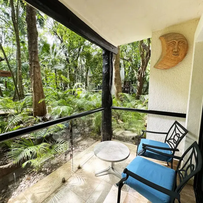 A private balcony at The Reef Playacar with two chairs, a table, and a lush green garden view surrounded by trees and plants.