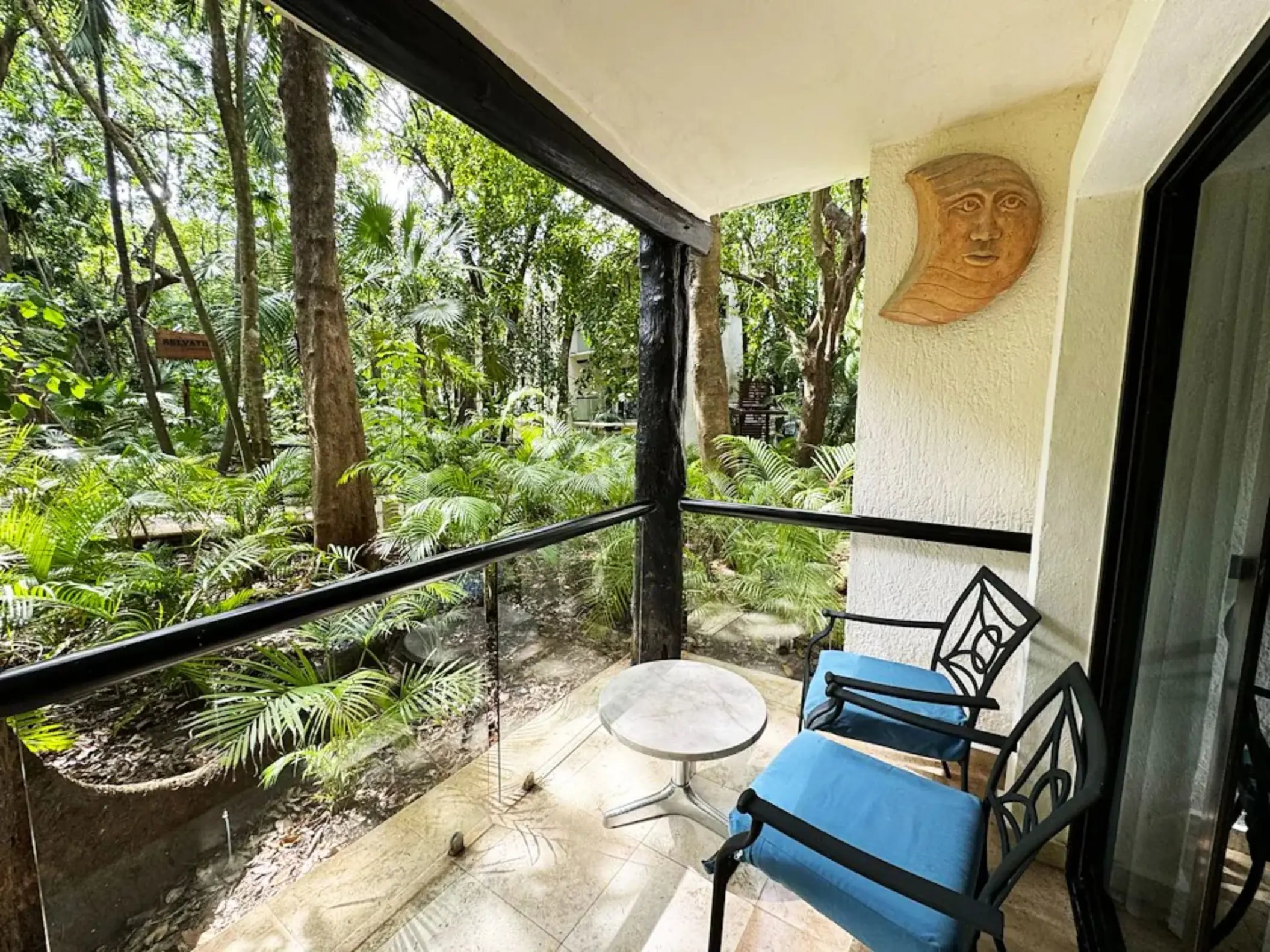 A private balcony at The Reef Playacar with two chairs, a table, and a lush green garden view surrounded by trees and plants.