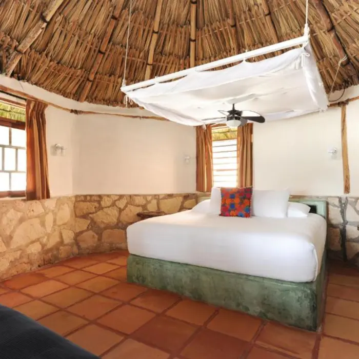 A rustic hotel room with a palm-thatched roof, stone walls, tiled floor, and a large white bed.