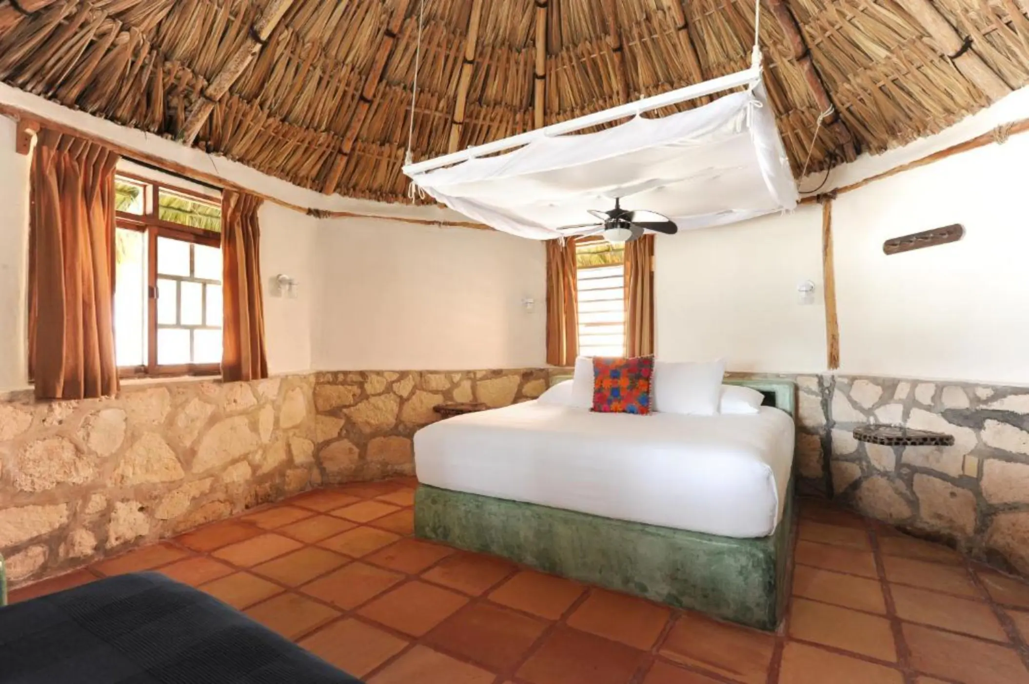 A rustic hotel room with a palm-thatched roof, stone walls, tiled floor, and a large white bed.