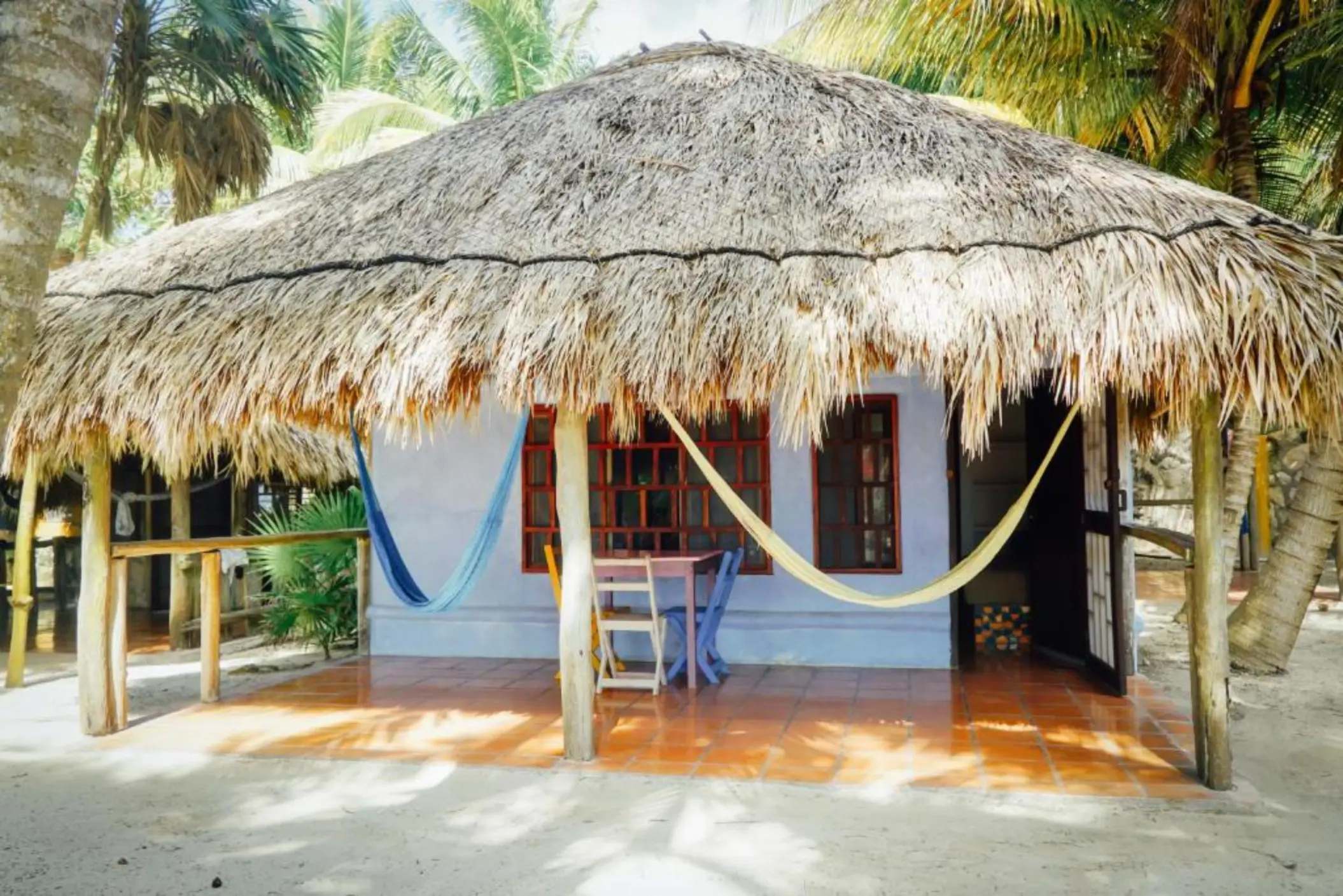 A rustic villa exterior with a thatched roof, blue walls, a porch with hammocks, and palm trees.