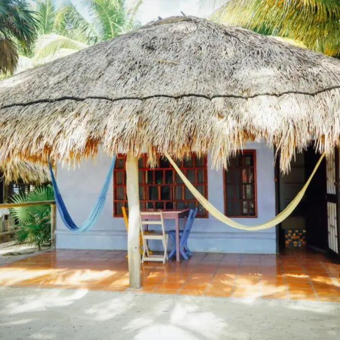 A rustic villa exterior with a thatched roof, blue walls, a porch with hammocks, and palm trees.