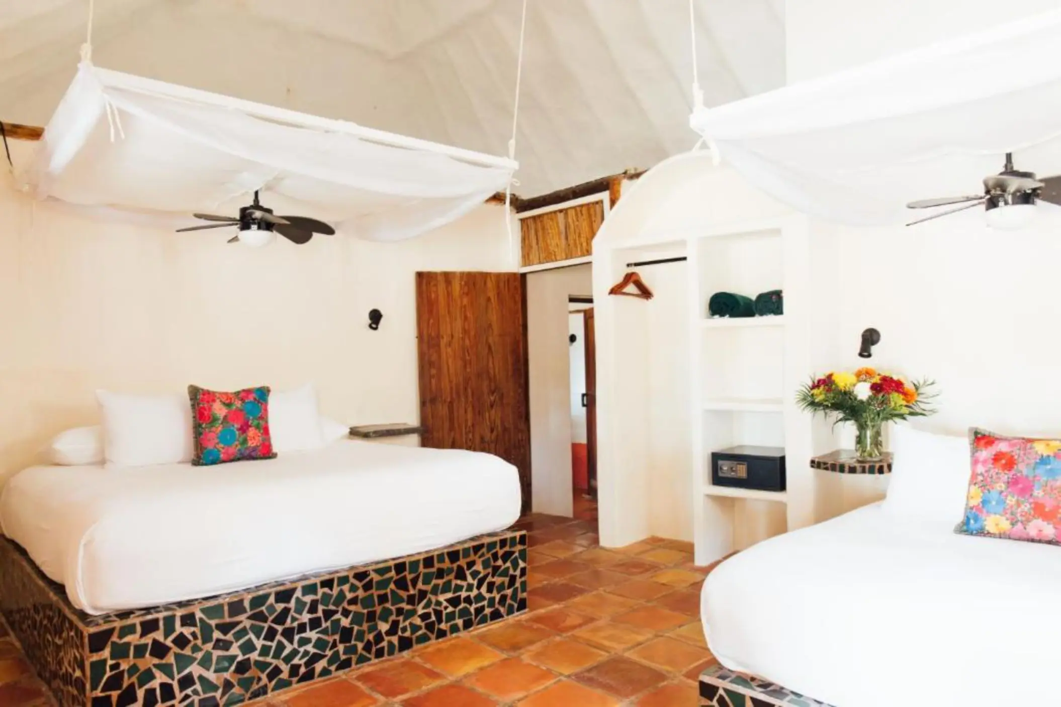 A hotel room with two beds with mosaic tile bases, a thatched roof, and white walls.
