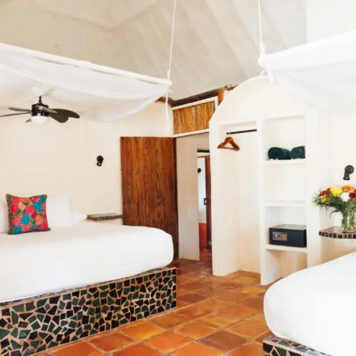 A hotel room with two beds with mosaic tile bases, a thatched roof, and white walls.