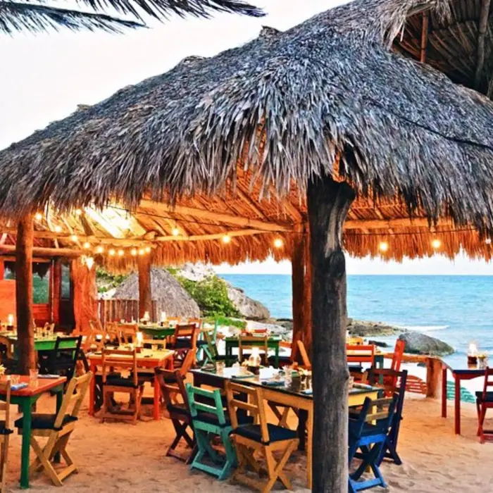 An oceanfront restaurant with a thatched roof, colorful wooden tables and chairs, and string lights.
