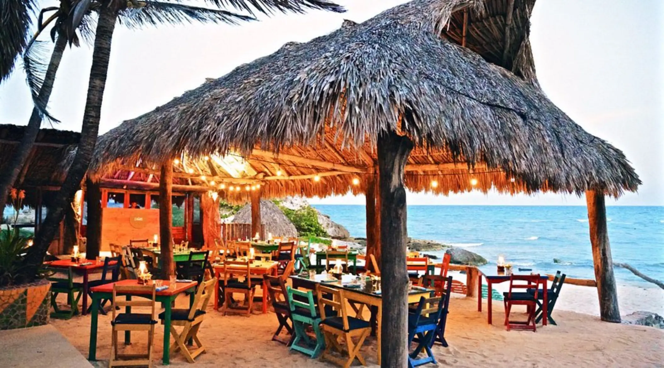 An oceanfront restaurant with a thatched roof, colorful wooden tables and chairs, and string lights.