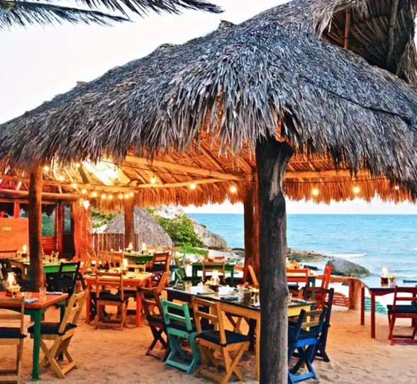 An oceanfront restaurant with a thatched roof, colorful wooden tables and chairs, and string lights.