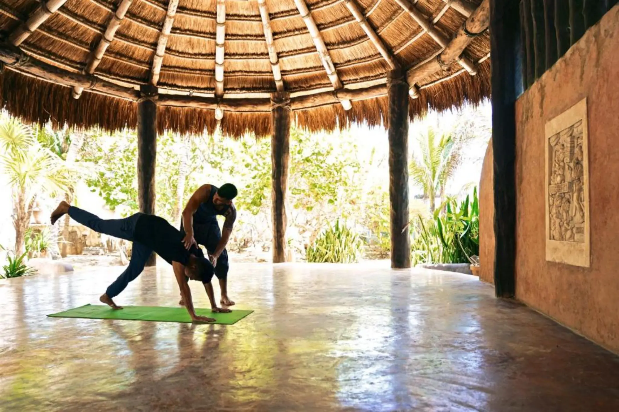 Two people doing yoga under a large, round thatched roof in a tropical setting.