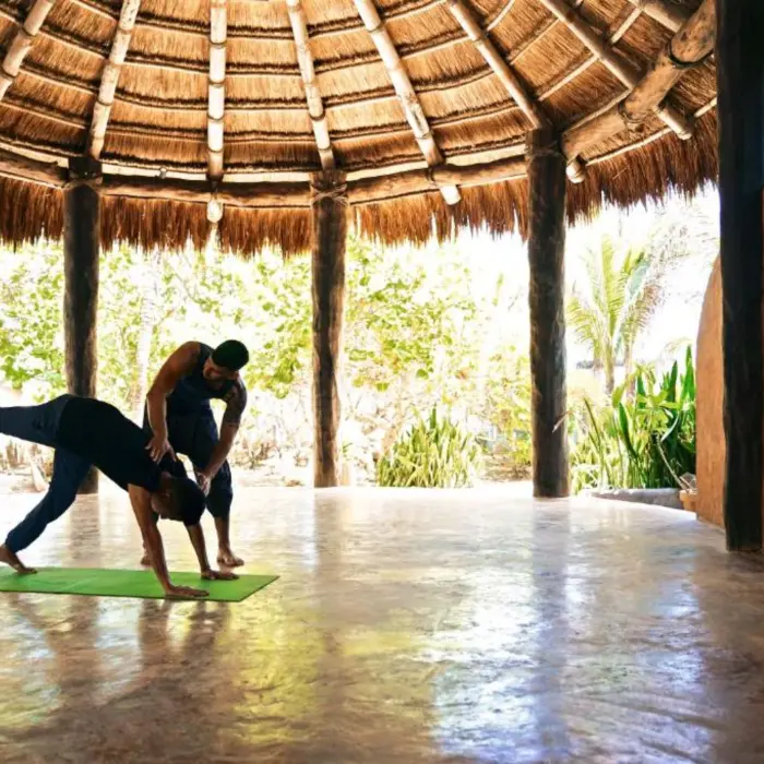 Two people doing yoga under a large, round thatched roof in a tropical setting.