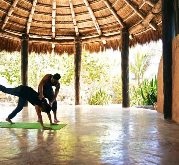 Two people doing yoga under a large, round thatched roof in a tropical setting.