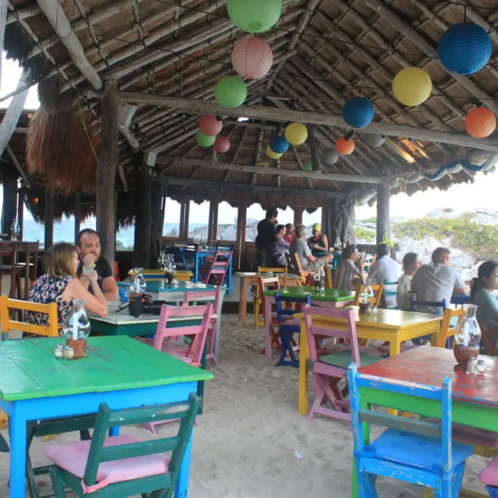 A colorful, rustic outdoor restaurant with a thatched roof, paper lanterns, and people dining.