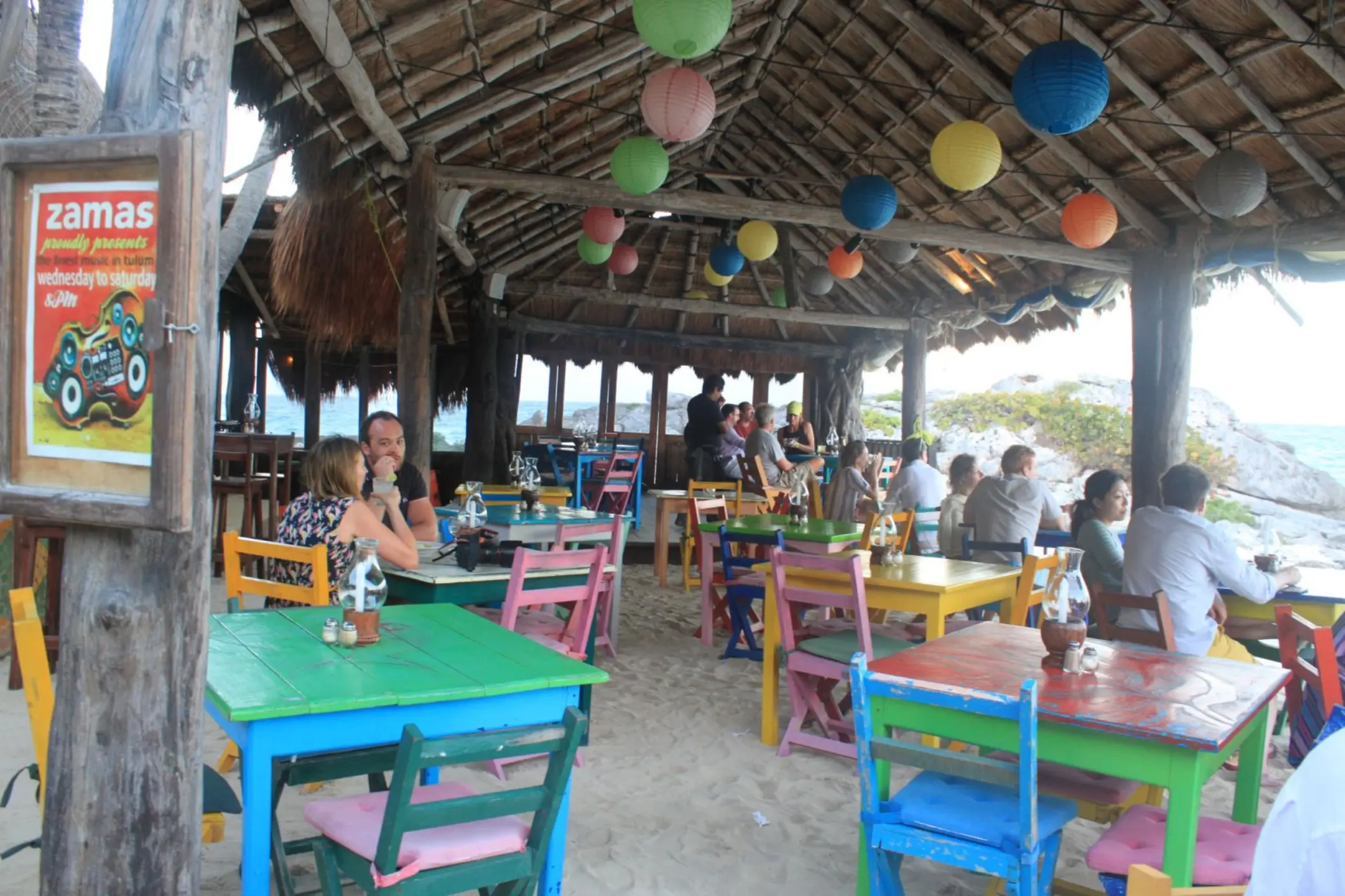 A colorful, rustic outdoor restaurant with a thatched roof, paper lanterns, and people dining.