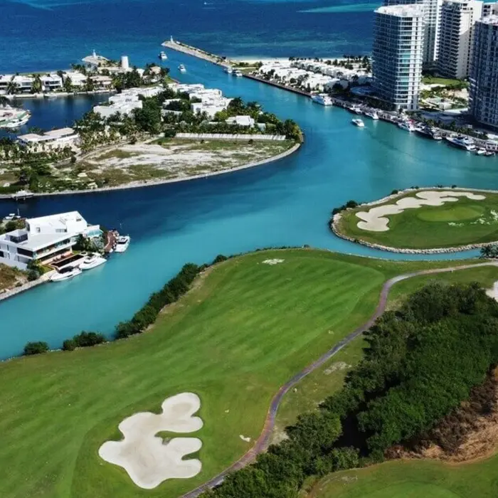 An aerial view of an island golf green with a winding sand bunker located directly next to the luxury marina and docked yachts in Puerto Cancún