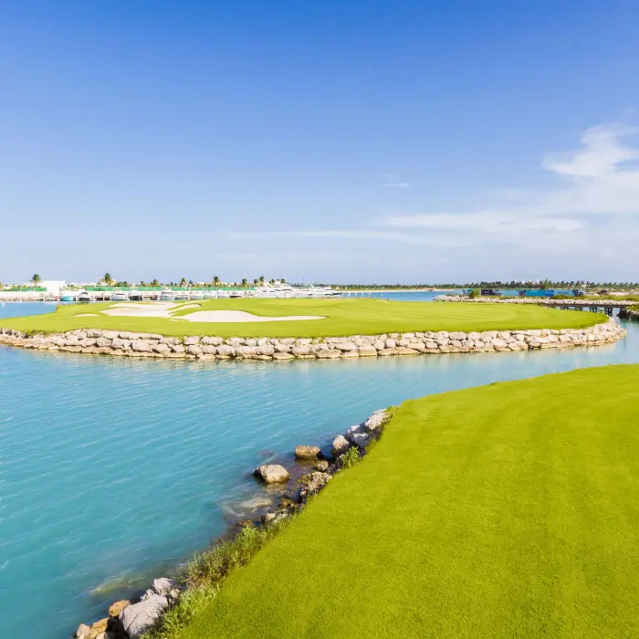 A vibrant golf green surrounded by bright turquoise water and stone walls, with the luxury Moon Palace resort towers in the distance